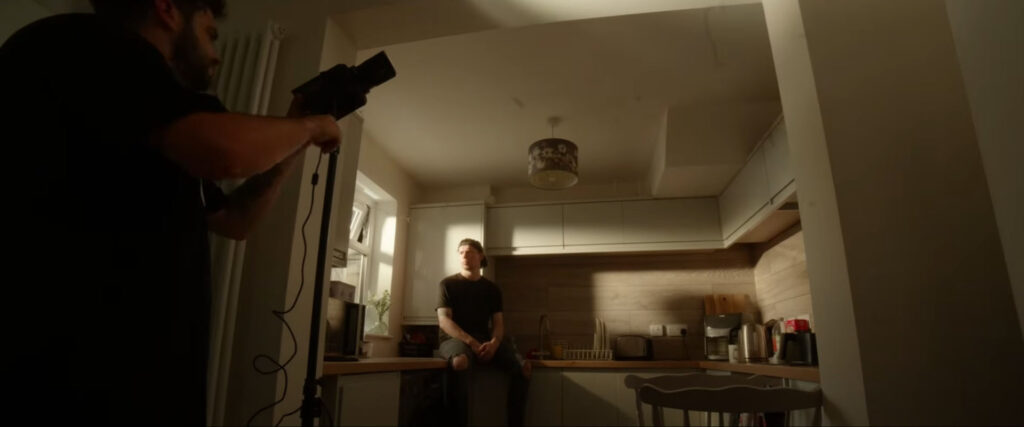 A man holding a light stands in a kitchen, while another man sits on a countertop bathed in sunlight.